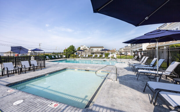 Outdoor pool area at Beachside Resort featuring a large swimming pool and a smaller shallow pool with metal handrails. Surrounding the pools are lounge chairs, blue umbrellas, and fenced green spaces with neighboring buildings in the background.