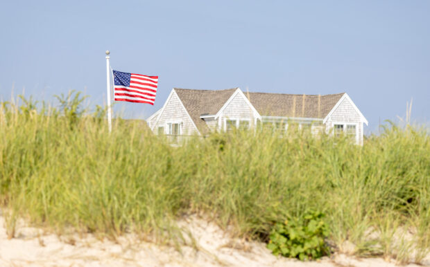 Close-up of an American flag waving in front of Beachside Village Resort, framed by tall dune grasses.