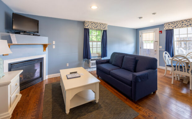 A cozy living room with a blue sofa, white coffee table, fireplace, and mounted TV. Navy curtains and patterned valances frame the windows, with a dining area featuring a white table and chairs nearby. Hardwood floors complete the space.