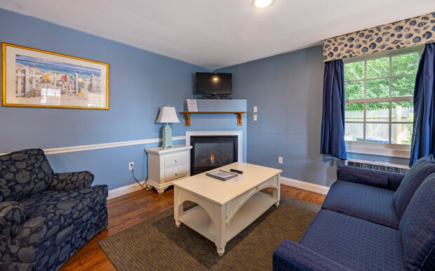 A cozy living room with a blue sofa, patterned armchair, fireplace with a mounted TV, and a white coffee table. Navy curtains frame a large window, and a beach-themed painting decorates the blue wall. Warm hardwood floors and a textured rug complete the space.