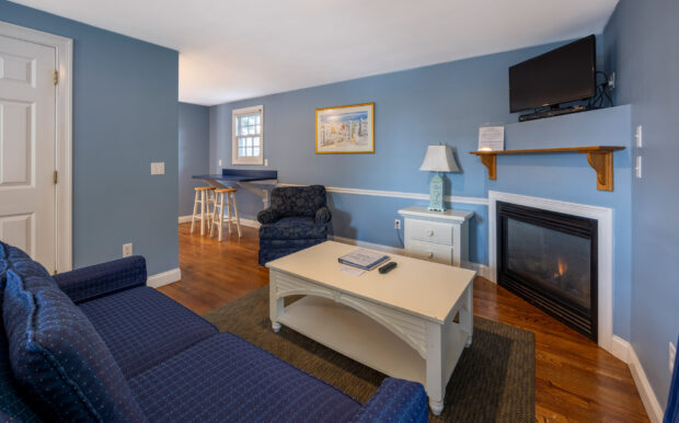 A cozy living room with a blue sofa, patterned armchair, white coffee table, and fireplace with a mounted TV. A small breakfast nook with stools sits in the corner. The soft blue walls feature a beach-themed painting, and hardwood floors add warmth to the space.