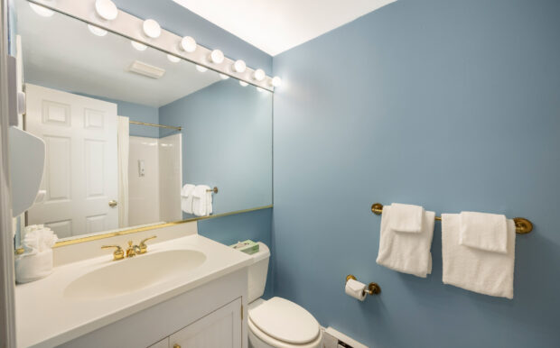 A bright bathroom with soft blue walls features a white vanity sink with gold fixtures, a large mirror with vanity lights, and neatly folded white towels on a gold towel bar. A shower with a curtain and a toilet are reflected in the mirror, completing the clean and inviting space.