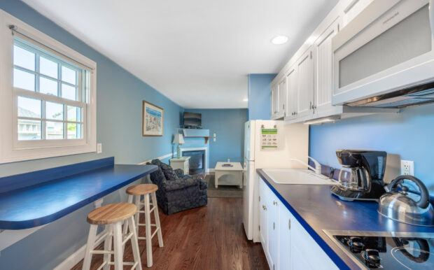 A cozy kitchen with white cabinetry and blue countertops features a stovetop, coffee maker, kettle, and microwave. A breakfast bar with two wooden stools sits by a window, and the space opens into a living area with a fireplace and TV. The blue walls and hardwood floors create a clean, inviting atmosphere.