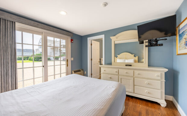 A bright bedroom with a large sliding glass door leading outside features a white bed, matching dresser with a mirror, and a mounted TV. Soft blue walls complement the hardwood floors and natural light, creating a calm and inviting atmosphere.