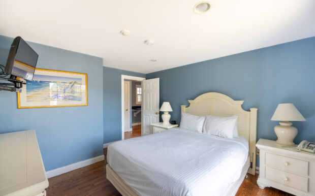 A serene bedroom with a white bed, matching headboard, and two nightstands featuring lamps. A mounted TV and a framed beach-themed painting adorn the soft blue walls, while hardwood floors add warmth to the space. An open doorway leads to an adjacent area.