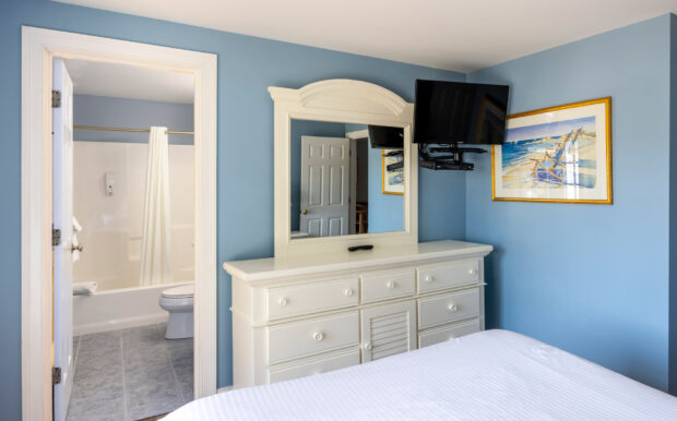 A bright bedroom features a white dresser with a matching mirror, a wall-mounted TV, and a beach-themed framed painting on soft blue walls. An open doorway leads to a bathroom with a bathtub, shower curtain, and white tiles, creating a clean and inviting space.