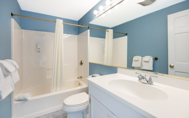 A clean, well-lit bathroom features a white bathtub with a shower curtain, a large mirror, and a white vanity with a sink. The blue walls are complemented by gold fixtures, neatly folded towels, and a bright vanity light fixture, creating a fresh and inviting space.