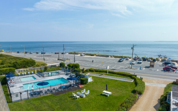 Aerial view of the pool area and green lawn at Beachside Village Resort, with picnic tables, lounge chairs, and the ocean in the background. A parking lot and road separate the resort from the beach.