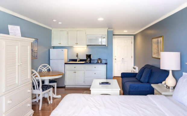 A welcoming studio featuring a kitchenette with white cabinets, navy blue countertops, and essential appliances. The dining table and seating area complement the cozy bed and blue-walled ambiance.