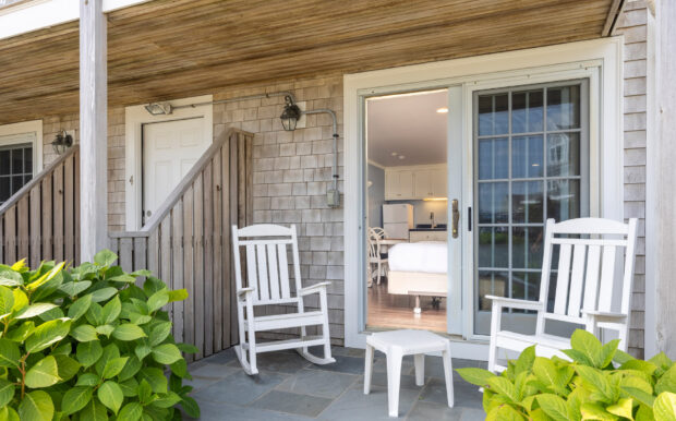 An outdoor patio area with white rocking chairs and a table. The space leads into the studio through sliding glass doors, surrounded by greenery for a serene atmosphere.