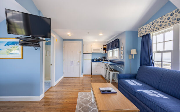 A bright studio room with a bed, kitchenette, navy sofa, and a white dresser. The space includes a skylight.