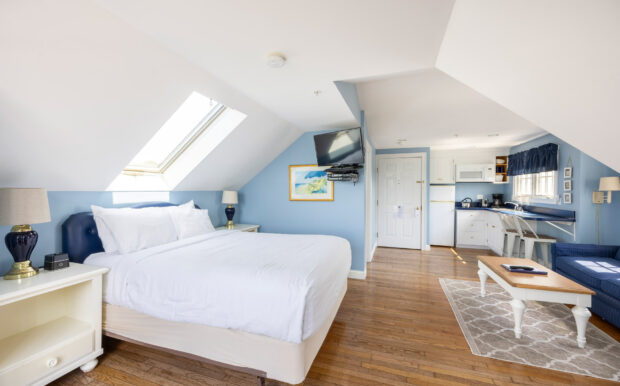 A studio bedroom with a white bed, nightstands, sloped ceilings, and a kitchenette in the corner.